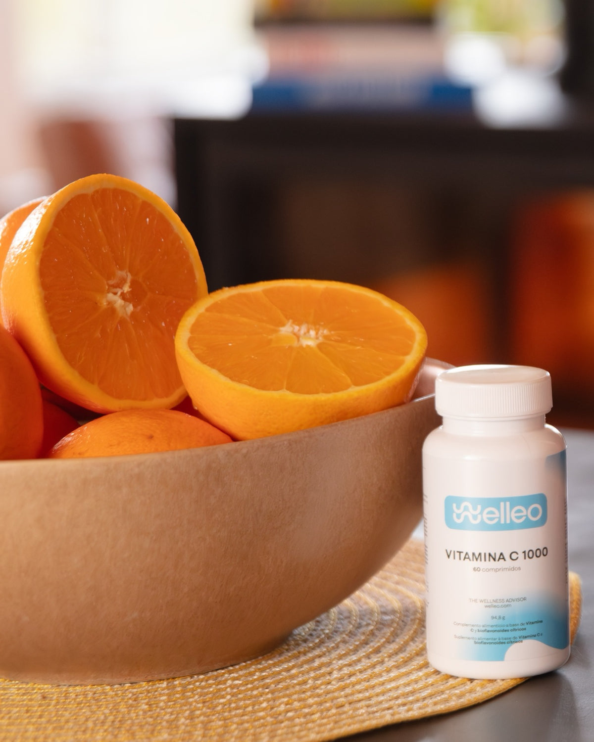 Un frasco de Vitamina C 1000 de welleo junto a un cuenco de naranjas partidas por la mitad